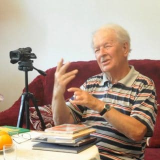 Pan Antoni Morawski udziela wywiadu, siedząc przy stole na którym leżą książki, obok ustawiona kamera