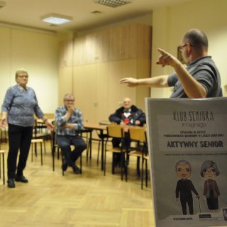 Plakat projektu Aktywny Senior, za nim w tle seniorzy w sali podczas warsztatów teatralnych