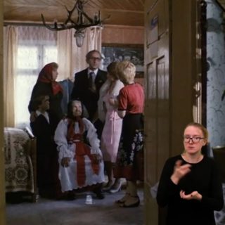 Kadr z filmu, rodzina stoi w pokoju wokół siedzącej babci ubranej odświętnie, w rogu ekranu tłumacz