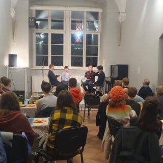 Studenci siedzący przy stolikach, przed nimi na scenie przedstawiciele fundacji fonis siedzą przy stoliku
