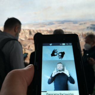 Ekran z przewodnikiem w języku migowym, w tle osoby oglądające Panoramę Racławicką