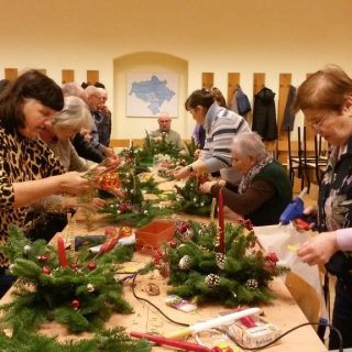 Grupa seniorów po obu stronach dużego stołu przygotowuje stroiki świąteczne