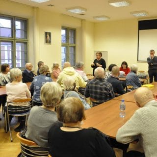 Seniorzy siedzą w sali wokół dwóch dużych stołów i patrzą na migającą panią Elę