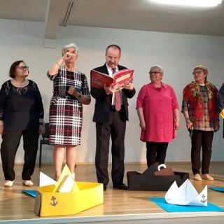 Grupa seniorów na scenie, na środku mężczyzna z otawrtą księgą, obok migająca kobieta