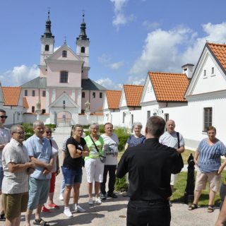 Grupa podcza zwiedzania patrzy na tłumacza języka migowego, w tle kościół na Wigrach