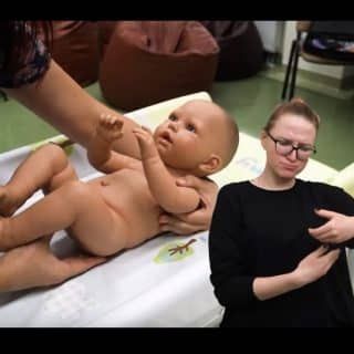 Ręcę chwytają dziecko (lalkę), obok tłumaczka języka migowego