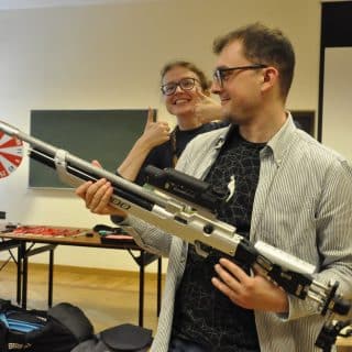 Mężczyzna z karabinkiem, zza niego wychyla się młoda kobieta z dwoma kcuikami skierowanymi do góry