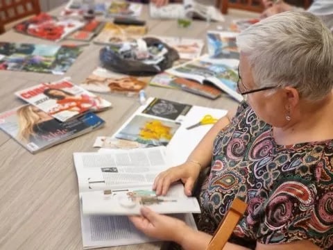 Warsztaty arteterapeutyczne Kobieta z otwartą książką przy stole na którym leżą gazety i inne rzeczy