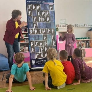 Przy tablicy z alfabetem migowym, mężczyzna uczy dziewczynkę migać, siedzące obok dzieci patrzą na nich
