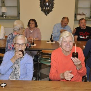 Dwie seniorki siedzące przy stołach pokazują naczynia, w tle seniorzy siedzą przy stołach