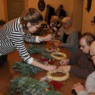 Młoda kobieta pomaga seniorom siedzącym przy stoliku tworzyć wieniec adwentowy
