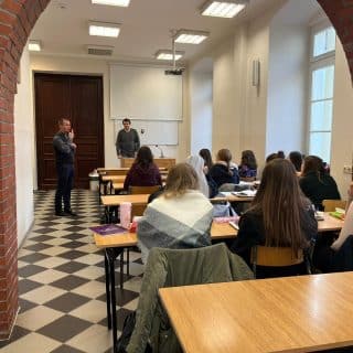 Widok z tyłu sali wykładowej na plecy studentów przed którymi przy katedrze stoi dwóch mężczyzn