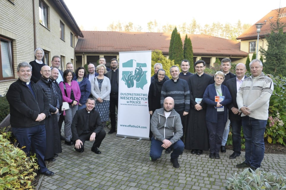 Zdjęcie grupowe. Grupa ludzi w ogrodzie, pośród nich baner Duszpasterstwa Niesłyszących w Polsce