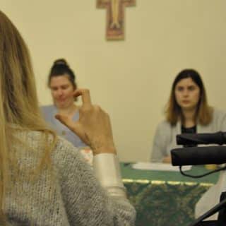 Dłoń migającej kobiety, w tle dwie inne kobiety siedzą przy stole