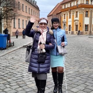 Osoba niewidoma z białą laską prowadzona pod rękę przez drugą kobietę