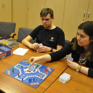 Dwie osoby z kartami siedzą przy stoliku z planszą i pionkami