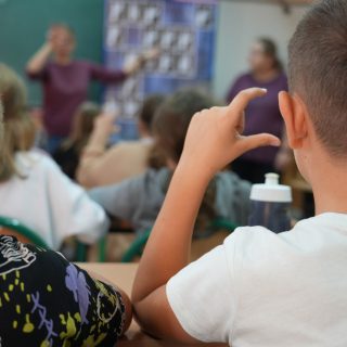 Chłopiec pokazuje literkę "C" z polskim alfabecie migowym, przed nim dzieci siedzące w ławkach i dwójka instruktorów