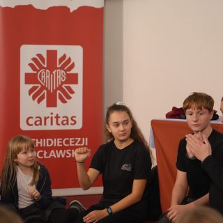 Grupa dzieci z instruktorami siedzi przy tablicy ze znakiem Caritas