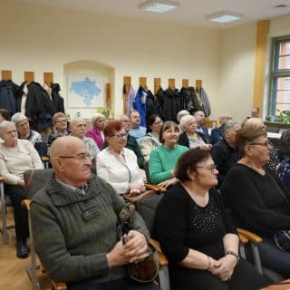 Grupa seniorów siedzi w sali w kilku rzędach