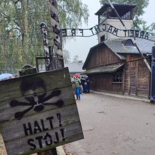 Drewniana tabliczka z trupią czaszką i napisem HALT! STÓJ! na tle bramy obozu w Oświęcimiu