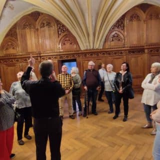 Grupa seniorów zwiedzające wnętrze sali ze ścianami obitymi drewnem i żebrowo-krzyżowym sklepieniem